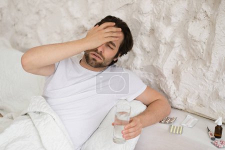 Foto de Despaired sad young caucasian man lies on bed with pills, suffers from headache, touch face with hand, holds glass of water in bedroom interior. Antidepressants and painkillers for migraine, emotions - Imagen libre de derechos