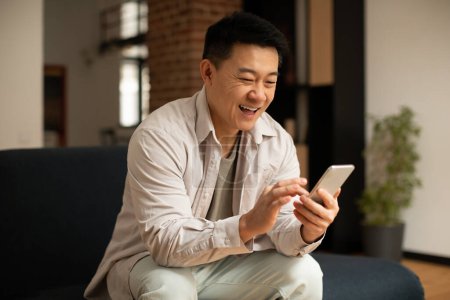 Téléchargez les photos : Asian middle aged man chatting on smartphone or surfing internet while resting on sofa in living room interior, copy space. Free time, new app, advertisement and offer - en image libre de droit