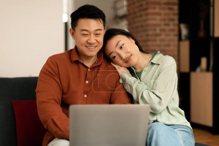 Téléchargez les photos : Loving asian couple using laptop together, surfing internet or watching movie online, wife leaning on husbands shoulder, sitting on sofa. Korean spouses using computer at home - en image libre de droit