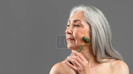 Téléchargez les photos : Serious old european lady with gray hair do jade massage, enjoy procedure with natural stone, isolated on gray background, studio, panorama. Facial massage for perfect skin, beauty care, spa treatment - en image libre de droit