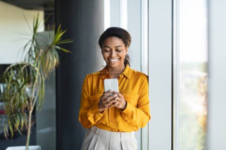 Photo for Happy successful black businesswoman typing on smartphone, standing near window in modern office interior, having chat with client online. Business at workplace, break from work - Royalty Free Image