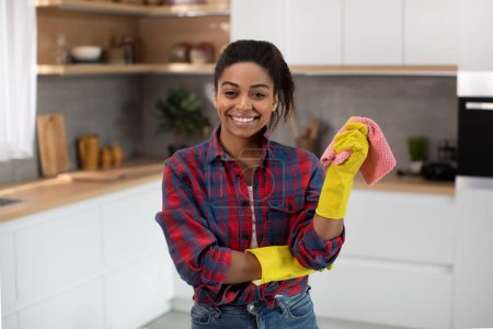 Photo for Cheerful pretty millennial african american housewife in rubber gloves with rag enjoy household chores in minimalist kitchen interior. Great cleaning service, hygiene at home, lifestyle, ad and offer - Royalty Free Image