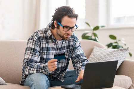Photo for Serious concentrated young caucasian male shopaholic in glasses sits on sofa with computer and credit card, shopping online, checks financial account in room interior. App banking, sale and money - Royalty Free Image