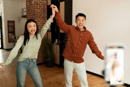 Photo for Couple of funny asian influencers shooting dance video for social network on cellphone, having fun at home. Man and woman bloggers streaming content for their vlog, broadcasting online - Royalty Free Image