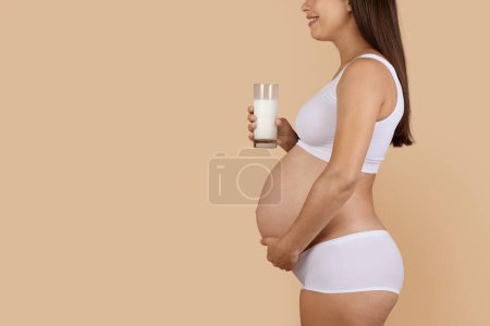 Photo for Dairy Foods In Pregnancy. Side View Shot Of Pregnant Woman Holding Glass Of Milk While Standing Isolated On Beige Background, Expectant Female In Underwear Embracing Belly And Enjoying Healthy Drink - Royalty Free Image