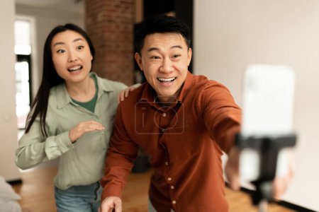 Photo for Excited asian man and woman bloggers broadcasting from home, sharing their lifestyle with followers, using smartphone on tripod, living room interior - Royalty Free Image