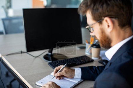 Photo for Online business class concept. Male office worker writing in notebook, taking notes and using computer with black screen for mockup template, watching lecture or webinar - Royalty Free Image