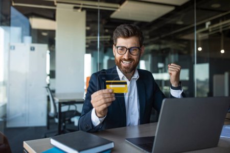 Téléchargez les photos : Excited middle aged businessman holding credit card and making YES gesture, sitting in front of laptop at workplace in office interior. Cool online promo concept - en image libre de droit