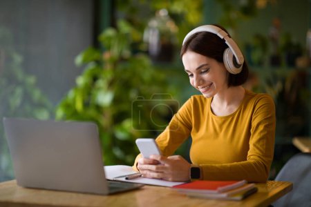 Photo for Cheerful young smiling IT woman using wireless headphones and phone, listen to music while work or study on laptop pc computer, sit at table in coffee shop cafe indoor. Freelance business concept - Royalty Free Image