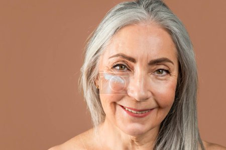 Photo for Happy old caucasian lady with natural beauty and cream on her face enjoy professional cosmetics, rejuvenation and spa treatments, isolated on brown background, studio. Skin care, anti-aging procedure - Royalty Free Image