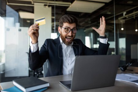 Foto de Overjoyed middle aged businessman with open mouth, screaming, holding credit card and looking at laptop in office interior. Successful business, trade, finance and win - Imagen libre de derechos