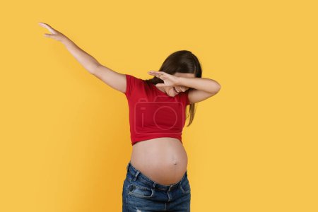 Foto de Young Pregnant Woman Throwing Dab Move And Laughing, Cheerful Millennial Expectant Lady Covering Face And Outstretching Hand, Dancing And Having Fun Over Yellow Studio Background, Copy Space - Imagen libre de derechos