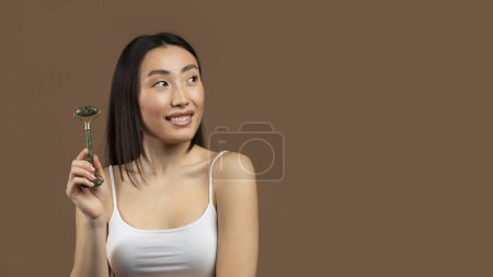 Téléchargez les photos : Pretty korean lady using greenstone jade facial roller, looking aside at free space while posing over brown studio background, panorama. Young asian woman caring for her skin - en image libre de droit