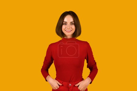 Photo for Cute pretty young brunette woman with nice hairstyle posing alone on orange studio background, holding hands in pockets, wearing red outfit, smiling at camera, copy space. Millennials lifestyle - Royalty Free Image