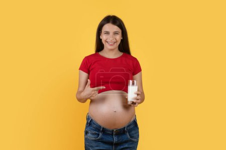 Foto de Beautiful Young Pregnant Woman Pointing At Glass Of Milk In Hand While Posing Over Yellow Background, Happy Expectant Female Recommending Healthy Calcium Drink And Smiling At Camera, Copy Space - Imagen libre de derechos