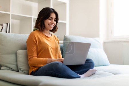 Photo for Online Communication. Portrait Of Beautiful Young Arab Woman Using Laptop At Home, Smiling Middle Eastern Female Sitting On Couch In Living Room Interior And Typing On Computer, Copy Space - Royalty Free Image