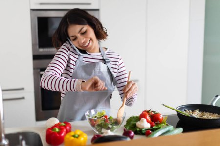 Téléchargez les photos : Cheerful young brunette pretty middle eastern woman wearing apron having phone conversation, looking at watch on her wrist, checking time while cooking dinner at home, waiting for guests, copy space - en image libre de droit