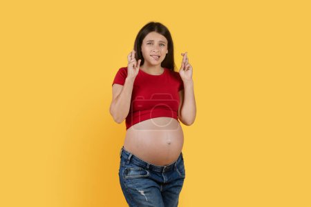Foto de Wxcited Pregnant Woman Making Wish With Crossed Fingers Over Yellow Studio Background, Superstitious Expectant Female Hoping For Luck And Healthy Pregnancy, Biting Lip And Looking At Camera - Imagen libre de derechos
