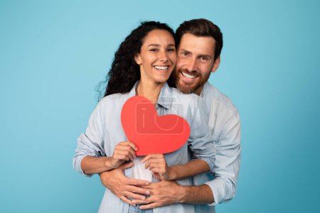 Téléchargez les photos : Smiling young european male in casual hug woman and holding heart, isolated on blue background, studio. Couple have fun, enjoy romantic, love and relationships, valentine day, anniversary celebration - en image libre de droit