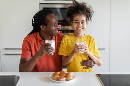 Foto de Happy black girl eating snacks in kitchen with her father, cheerful african american family dad and preteen daughter enjoying homemade muffins and drinking milk, having fun together at home - Imagen libre de derechos
