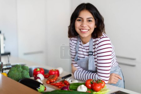 Photo for Portrait of cheerful attractive arabic young woman wearing grey apron chef posing at cozy kitchen at home, lady cooking healthy delicious food, using only eco organic vegetables, copy space - Royalty Free Image