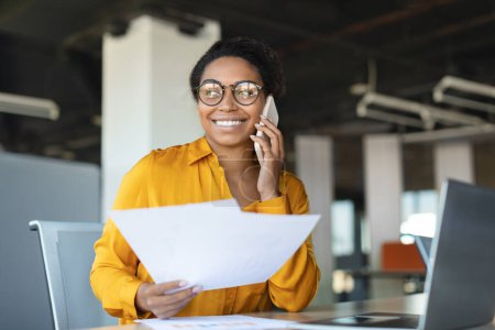 Foto de Happy african american businesswoman sitting at workdesk, having phone call with business partner, holding papers and smiling, working in office, copy space - Imagen libre de derechos