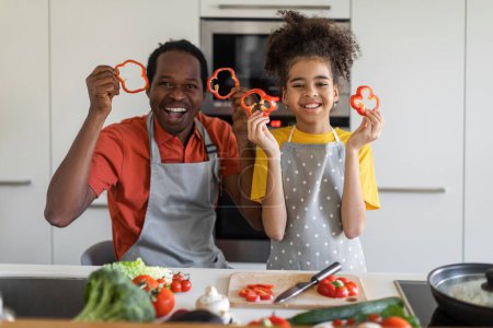Téléchargez les photos : Portrait Of Cheerful Black Father And Daughter Holding Bell Pepper Slices And Smiling At Camera While Posing In Kitchen, Happy African American Dad And Female Child Having Fun While Cooking At Home - en image libre de droit