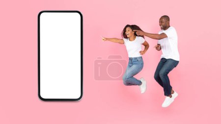 Téléchargez les photos : Overjoyed african american spouses jumping in air, pointing fingers at cellphone with blank screen, pink background, mockup. Surprise online, win, great ad, offer and news, app for fun together - en image libre de droit
