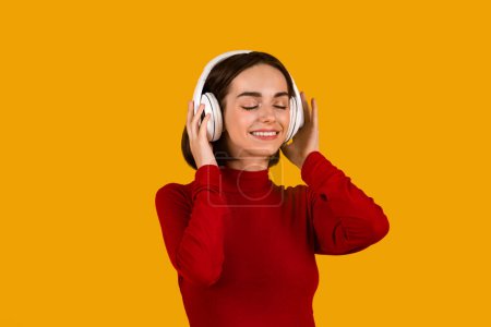 Foto de Young pretty smiling brunette lady in red longsleeve listening to music with closed eyes, using modern wireless stereo headphones and enjoying sound, isolated over orange studio background, closeup - Imagen libre de derechos