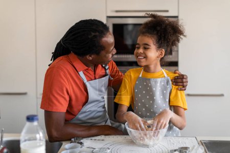 Foto de Happy black dad and daughter baking pastry together at kitchen, cheerful african american father and preteen female child making dough for cookies, enjoying cooking at home, closeup portrait - Imagen libre de derechos