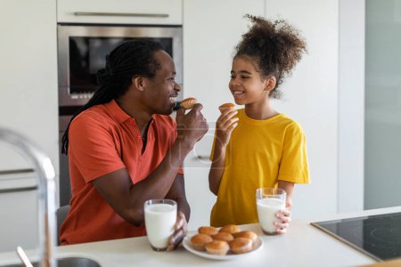 Téléchargez les photos : Portrait Of Cute Preteen Black Girl Eating Snacks With Dad In Kitchen, Happy African American Father And Daughter Enjoying Tasty Muffins And Drinking Milk While Having A Bite Together At Home - en image libre de droit