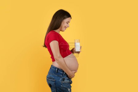 Foto de Pregnancy Diet. Smiling Young Woman Expecting Baby Holding Glass Of Milk While Standing Isolated Over Yellow Background, Side View Shot Of Happy Pregnant Female Enjoying Healthy Calcium Drink - Imagen libre de derechos