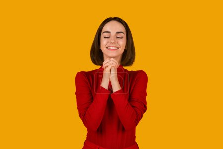 Photo for Merry cute pretty young lady with nice hairstyle in red outfit praying with closed eyes and smiling on orange studio background, woman ask for dreams come true, bright future, make a wish, copy space - Royalty Free Image