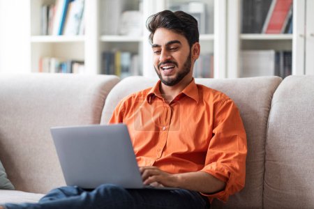 Téléchargez les photos : Happy attractive bearded young arabic man freelancer working from home, middle eastern guy sitting on couch in living room, using laptop, programming, copy space. Remote job concept - en image libre de droit