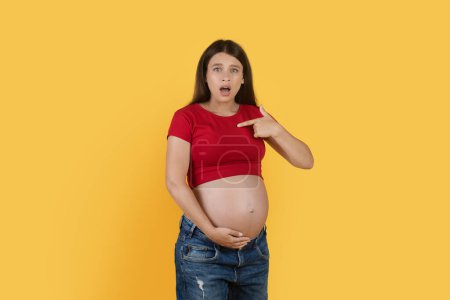Téléchargez les photos : Why Me. Confused Young Pregnant Woman Pointing At Herself And Looking At Camera, Shocked Expectant Female Embracing Her Big Belly While Standing Isolated Over Yellow Background, Copy Space - en image libre de droit