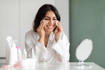 Foto de Pretty smiling young middle eastern lady in white silk bathrobe sitting at vanity table full of cosmetics at home, looking at mirror, using cotton pads, removing makeup before sleep, copy space - Imagen libre de derechos