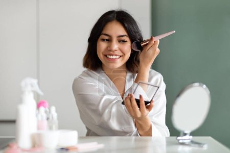 Téléchargez les photos : Ideas for everyday makeup. Beautiful cheerful middle eastern young woman in white silk bathrobe sitting at vanity table at home, looking at mirror and applying brown blush on her cheeks, smiling - en image libre de droit