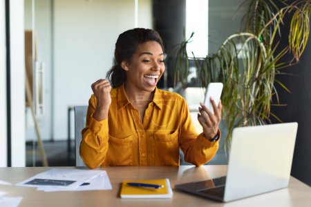 Foto de Excited black woman reading good message on smartphone, working on laptop in modern office interior. Great news, successful business, development, planning and financial strategies with online app - Imagen libre de derechos