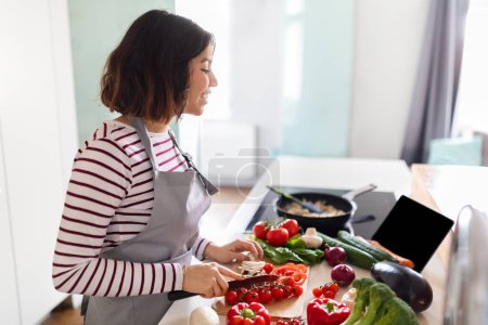 Photo for Side view of pretty young woman cooking delicious healthy food at home, using digital tablet with black empty screen, checking nice cooking application on pad, watching food vlog, copy space, mockup - Royalty Free Image