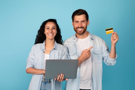 Photo for Glad young european man and arab woman with laptop show finger on credit card, recommend banking, isolated on blue background, studio. Advice for online shopping for shopaholics, sale and cashback - Royalty Free Image