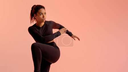 Photo for Fitness black woman standing doing elbow to knee crunch, looking aside at free space over neon peach studio background, panorama. Fitness workout, bodyshaping - Royalty Free Image