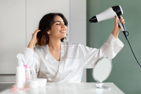 Téléchargez les photos : Happy attractive brunette young arabic woman enjoying hair drying routine, cheerful middle eastern lady in bathrobe styling her short hair with fan, sitting at vanity table, home interior - en image libre de droit