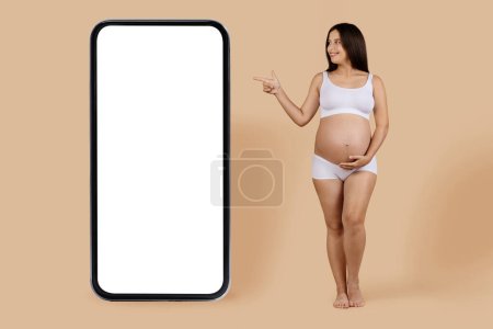 Téléchargez les photos : Online Ad. Happy Young Pregnant Female Pointing Aside At Big Blank Smartphone With White Screen, Smiling Expectant Woman In Underwear Demonstrating Copy Space,Standing On Beige Background, Mockup - en image libre de droit