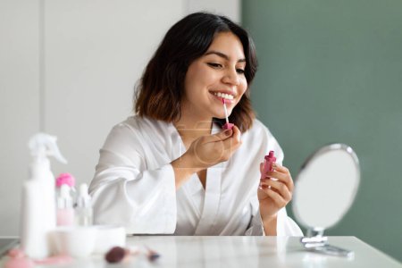 Photo for Beautiful young middle eastern woman looking at mirror, applying lipstick, home interior, empty space. Attractive lady taking care of her beautiful lips, putting bright lip gloss, applying makeup - Royalty Free Image