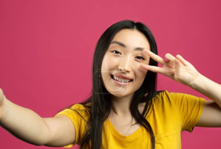 Photo for Portrait of joyful chinese lady taking selfie picture, point of view pov shot of happy female looking at camera and showing v victory sign peace gesture, pink background - Royalty Free Image