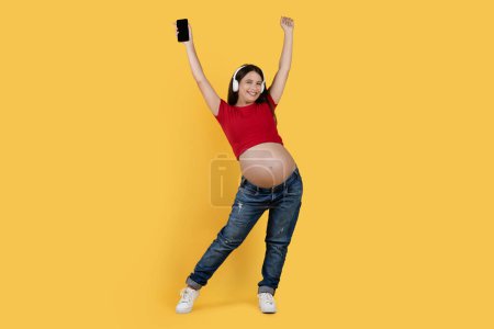 Foto de Cheerful Pregnant Female In Headphones Holding Smartphone And Dancing, Happy Young Expectant Woman Listening Music Online, Having Fun While Posing Over Yellow Studio Background, Copy Space - Imagen libre de derechos