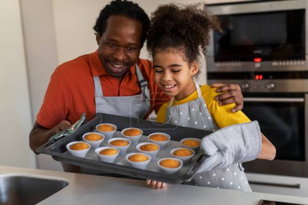 Foto de Cute black girl and her dad holding tray with muffins in kitchen, excited african american father and preteen female child looking at fresh baked pastry, enjoying cooking together at home - Imagen libre de derechos