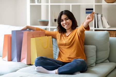 Foto de Happy Young Arab Woman Sitting On Couch With Shopping Bags And Showing Credit Card, Smiling Middle Eastern Female Enjoying Seasonal Sales And Discounts, Recommending Easy Payments, Free Space - Imagen libre de derechos