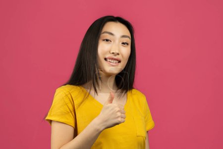Téléchargez les photos : Best choice concept. Cheerful asian young lady showing thumb up gesture, recommending or approving something, giving positive feedback and smiling on pink studio background - en image libre de droit