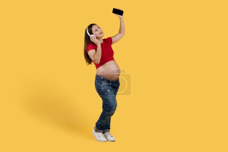 Foto de Positive Pregnant Woman In Headphones Listening Music On Smartphone And Singing, Cheerful Young Expectant Female Having Fun While Standing Over Yellow Studio Background, Full Length, Copy Space - Imagen libre de derechos